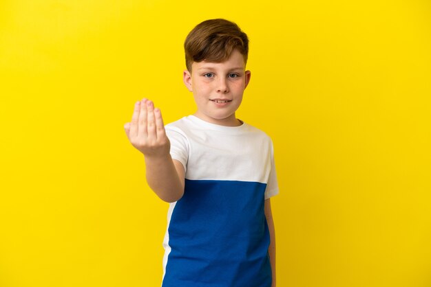 Niño pelirrojo aislado sobre fondo amarillo que invita a venir con la mano. Feliz de que hayas venido