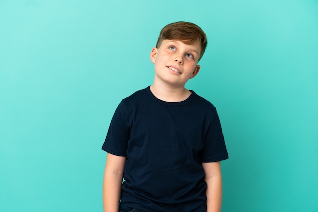 Niño pelirrojo aislado sobre fondo azul pensando en una idea mientras mira hacia arriba