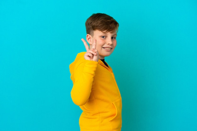 Niño pelirrojo aislado sobre fondo azul sonriendo y mostrando el signo de la victoria