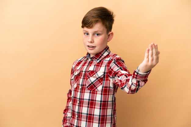 Niño pelirrojo aislado sobre fondo beige que invita a venir con la mano. Feliz de que hayas venido