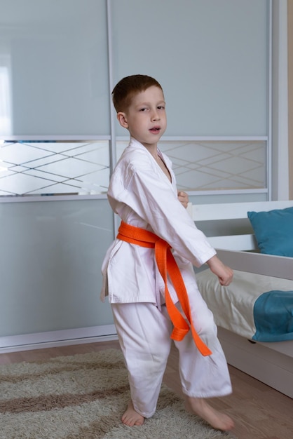 Niño pelirrojo se dedica a patadas de entrenamiento de karate en casa en forma de kimono con un deporte infantil de cinturón naranja