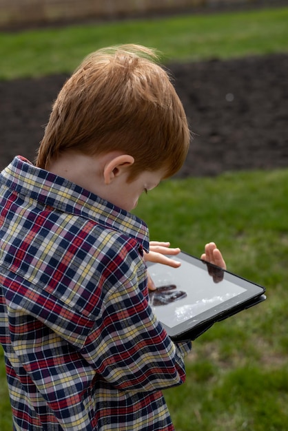 Niño pelirrojo feliz jugando en una tableta o viendo dibujos animados caminando en el patio trasero en la vista de las vacaciones de verano del pueblo desde atrás