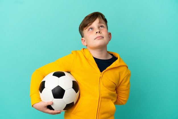 Niño pelirrojo jugando al fútbol aislado sobre fondo azul y mirando hacia arriba