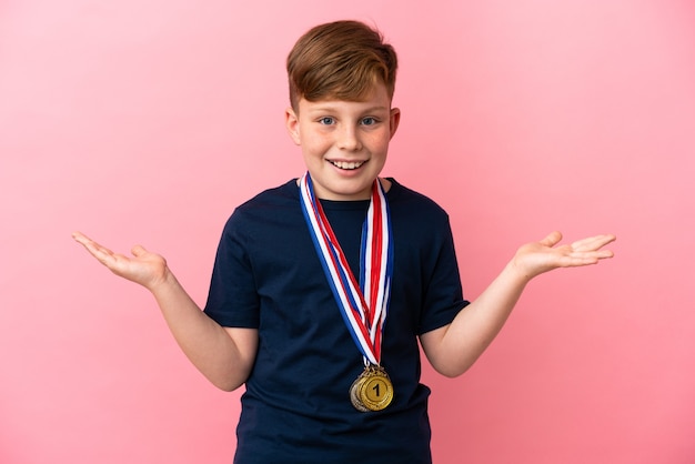 Niño pelirrojo con medallas aislado sobre fondo rosa con expresión facial sorprendida