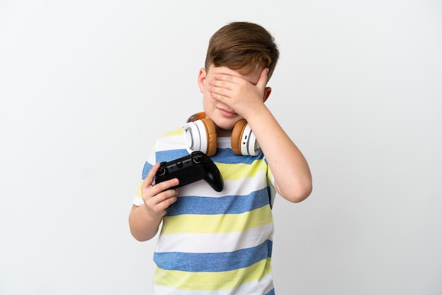 Niño pelirrojo sosteniendo un mando de juego aislado en la superficie blanca que cubre los ojos con las manos. No quiero ver algo