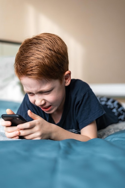 Niño pelirrojo sostiene un teléfono móvil en sus manos mira dibujos animados y videojuegos El retrato de estilo de vida se encuentra en la cama en casa y sonríe fotografía vertical