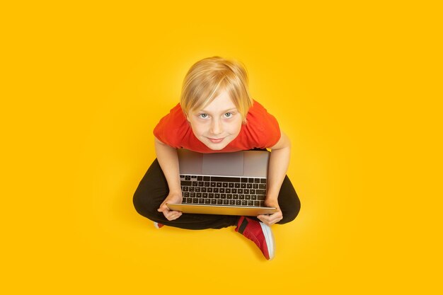 Niño de pelo blanco se sienta en el piso con una computadora portátil y mira a la cámara Retrato de vista superior de un niño con una computadora sobre fondo amarillo