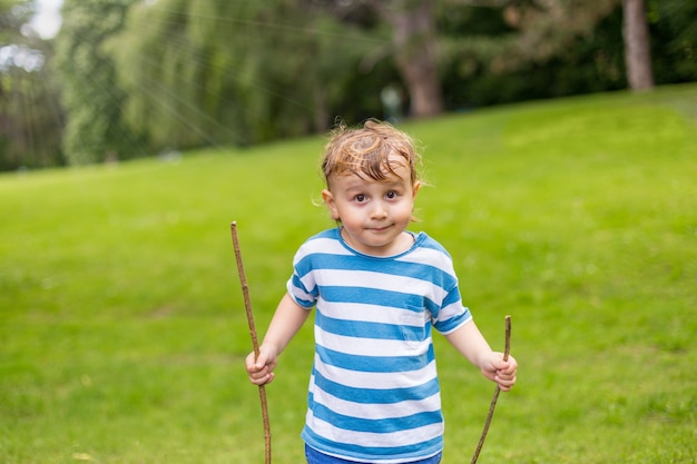 Niño con pelos sudorosos sosteniendo palos de madera y jugando en el parque en verano