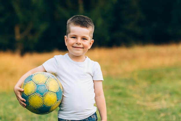 Foto niño con una pelota de fútbol bajo el brazo en el jardín de la casa con fondo de hierba, sonriendo