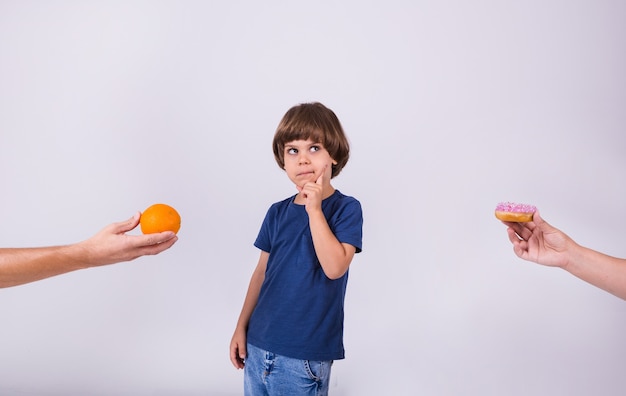 Un niño pensativo con una camiseta hace una elección entre una naranja y una rosquilla sobre un fondo blanco con un lugar para el texto