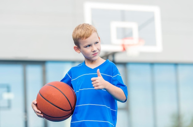 Niño pensativo gestando los pulgares hacia arriba mientras sostiene la pelota contra el aro de baloncesto