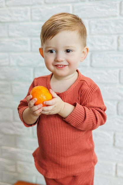 Un niño pequeño de 2 años sostiene un cítrico naranja en sus manos. El niño quiere comer mandarina.