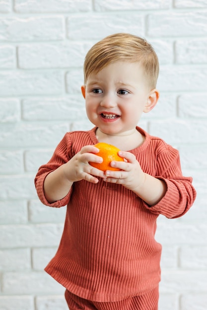 Un niño pequeño de 2 años sostiene un cítrico naranja en sus manos. El niño quiere comer mandarina.