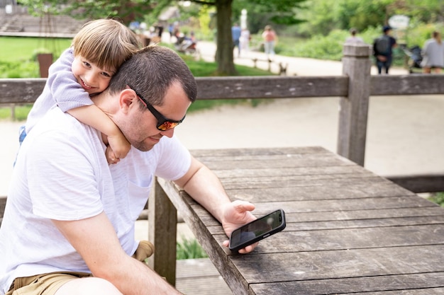 Niño pequeño abraza fuertemente a su padre mirando el teléfono inteligente en el parque en verano Niño molestando a su padre mientras usa internet y trabaja