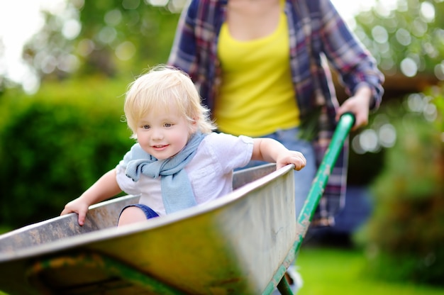 Niño pequeño adorable que se divierte en una carretilla que empuja por la mamá en jardín nacional, en día soleado caliente. Juegos al aire libre activos para niños en verano.