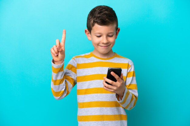 Foto niño pequeño aislado sobre fondo azul usando teléfono móvil y levantando el dedo