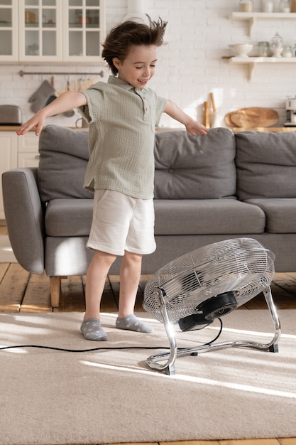 Un niño pequeño alegre solo en la sala de estar disfruta de la brisa fresca del ventilador o aire acondicionado retro