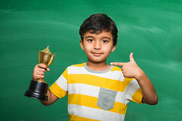 Niño pequeño asiático indio de la escuela que sostiene el trofeo o la copa ganadora contra la pizarra blanca o verde