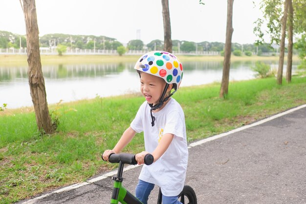 Niño pequeño asiático que usa el casco de seguridad aprendiendo a montar la bicicleta de primer equilibrio en un día soleado de verano