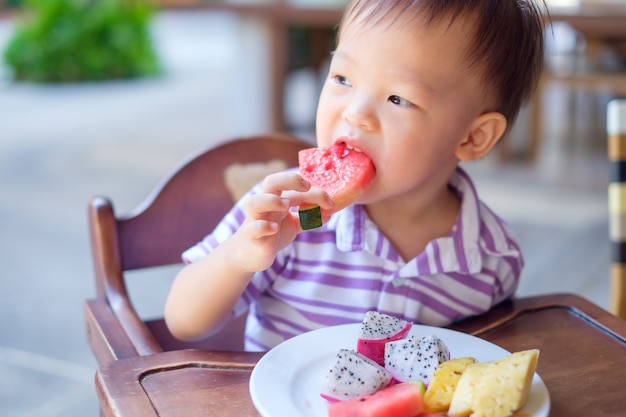 Niño pequeño asiático sentado en una silla alta con las manos comiendo sandía, piña, fruta del dragón