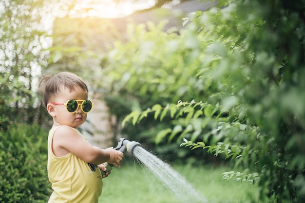 Niño pequeño asiático vertiendo agua en los árboles. El niño ayuda a cuidar las plantas con una regadera en el jardín.