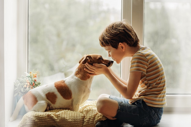 El niño pequeño besa al perro en nariz en la ventana.