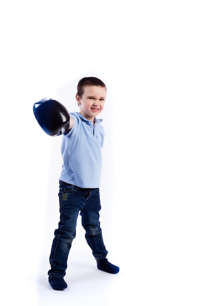 Un niño pequeño con cabello oscuro en jeans azules, una camisa de polo azul con guantes de boxeo blancos y negros se divierte boxeando y se considera un ganador en un fondo blanco aislado en un estudio fotográfico