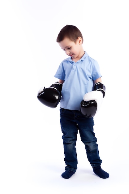 Un niño pequeño con cabello oscuro en jeans azules, una camisa de polo azul con guantes de boxeo blancos y negros se divierte boxeando y se considera un ganador en un fondo blanco aislado en un estudio fotográfico