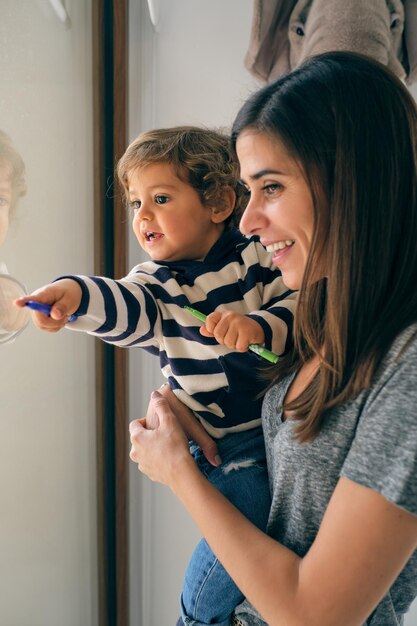 Niño pequeño de cabello rizado emocionado en suéter a rayas con marcadores apuntando cerca de la ventana con madre sonriente en ropa casual en casa