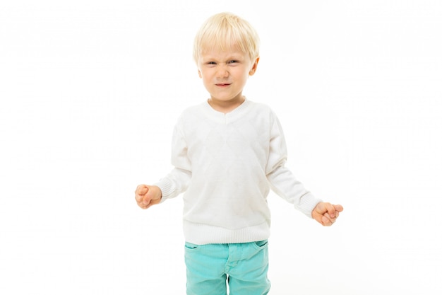 Niño pequeño con cabello rubio corto, ojos azules, apariencia linda, chaqueta blanca, pantalón azul claro, de pie y sonriente