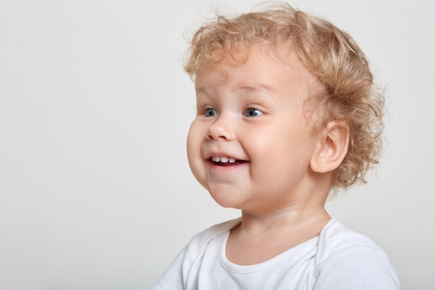 Niño pequeño con cabello rubio rizado riendo mientras mira atentamente a lo lejos mientras posa aislado sobre el espacio en blanco