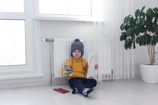 Foto un niño pequeño está cálidamente vestido con un suéter se sienta cerca de la batería con facturas impuestos y dinero