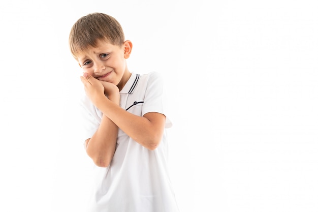 Un niño pequeño con una camisa blanca, pantalones cortos azules con cabello rubio, tiene las manos detrás de la mejilla, le duele el diente