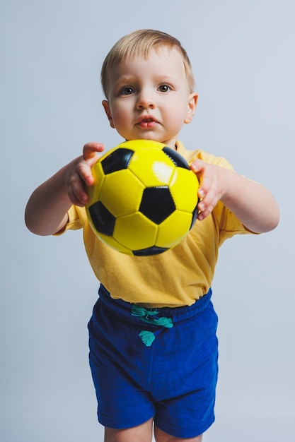 Un niño pequeño con una camiseta amarilla con una pelota de fútbol en la mano sonríe aislado en un fondo blanco Niño deportivo sosteniendo una pelota Juego de deportes para niños Pequeño atleta