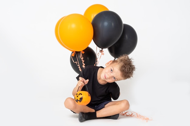 Un niño pequeño con una camiseta negra sosteniendo una canasta para golosinas y globos naranjas y negros asusta sentado en un fondo blanco