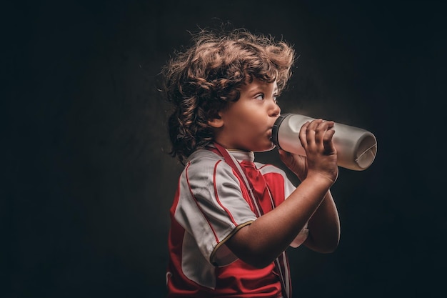 Niño pequeño campeón en ropa deportiva con una medalla de oro bebiendo agua de una botella. Aislado en el fondo oscuro con textura.