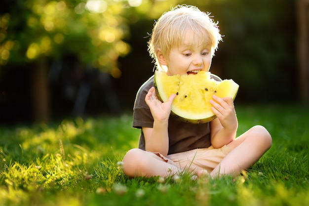 Niño pequeño caucásico lindo con los pelos rubios que come la sandía amarilla al aire libre