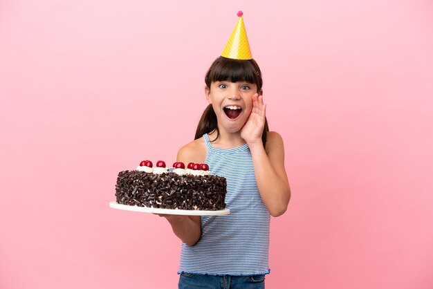 Niño pequeño caucásico sosteniendo pastel de cumpleaños aislado en fondo rosa con sorpresa y expresión facial conmocionada