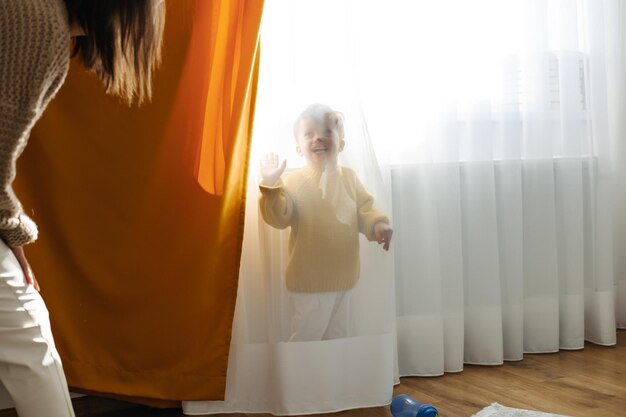 Un niño pequeño con una chaqueta amarilla se esconde de su madre detrás de una cortina en la guardería.