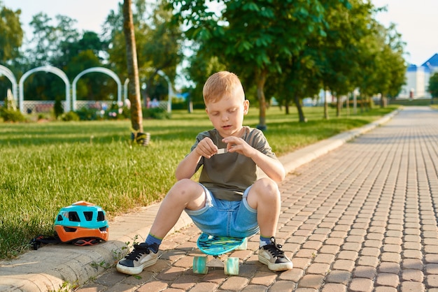 Un niño pequeño de cinco años está sentado en una patineta y quiere sellar la herida con una tirita.