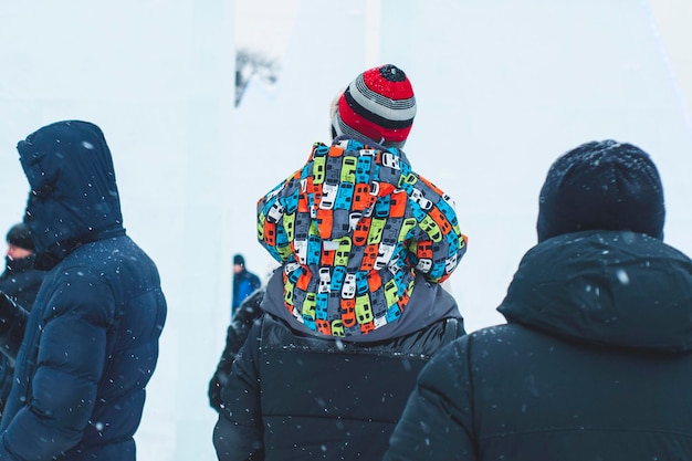 Un niño pequeño con una colorida chaqueta brillante se sienta sobre los hombros de su padre en el invierno en la calle