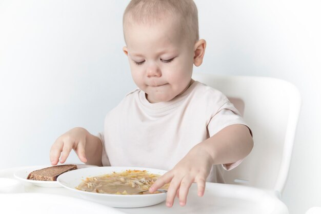 Un niño pequeño come sopa con pan solo en una trona contra el fondo de una pared blanca