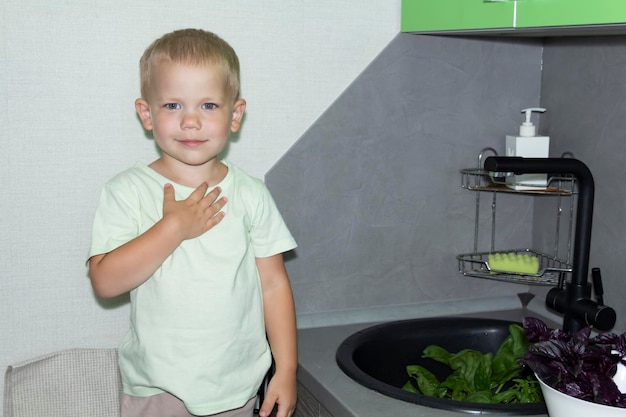Un niño pequeño con un corte de pelo corto ayuda a cocinar en la cocina Lava verduras y hierbas frescas en un fregadero negro en una cocina gris