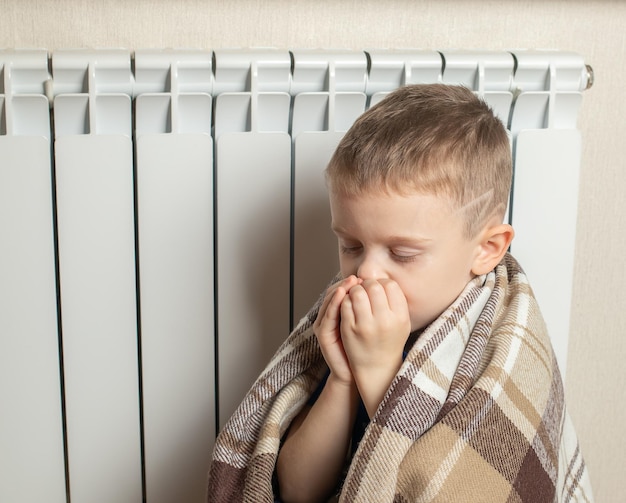 Un niño pequeño cubierto con una manta se está calentando en casa junto al radiador El comienzo de la temporada de calefacción El concepto de ahorro de calor Crisis energética