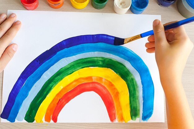 Foto un niño pequeño dibuja un arco iris con lápices de punta de fieltro en un cuaderno de bocetos mientras está sentado en una mesa