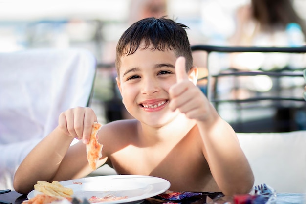 Niño pequeño disfrutando de comer después de salir de la piscina, mirando a la cámara y riendo