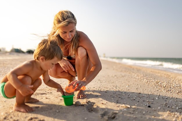 Un niño pequeño y divertido, recoge conchas y guijarros en el mar azul tranquilo en un fondo arenoso junto con una madre amable y cariñosa bajo el sol de verano en unas vacaciones brillantes