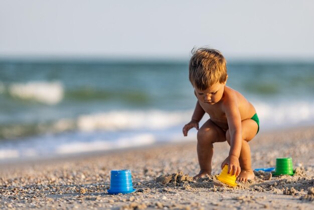 Un niño pequeño y divertido, recoge conchas y guijarros en el mar azul tranquilo en un fondo arenoso bajo el sol de verano en unas vacaciones brillantes