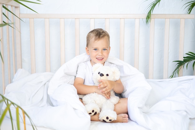 Un niño pequeño se encuentra en una cuna con un oso de peluche en la ropa de cama blanca un sueño infantil sonríe