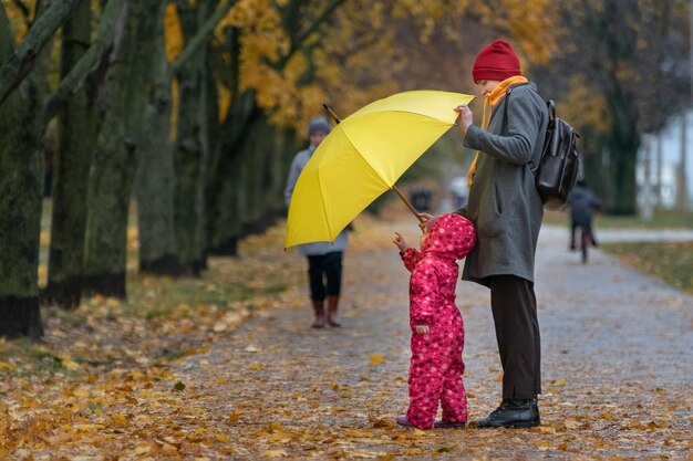 El niño pequeño se encuentra en la lluvia amarilla junto a su madre en el parque con el telón de fondo del follaje amarillo otoñal.